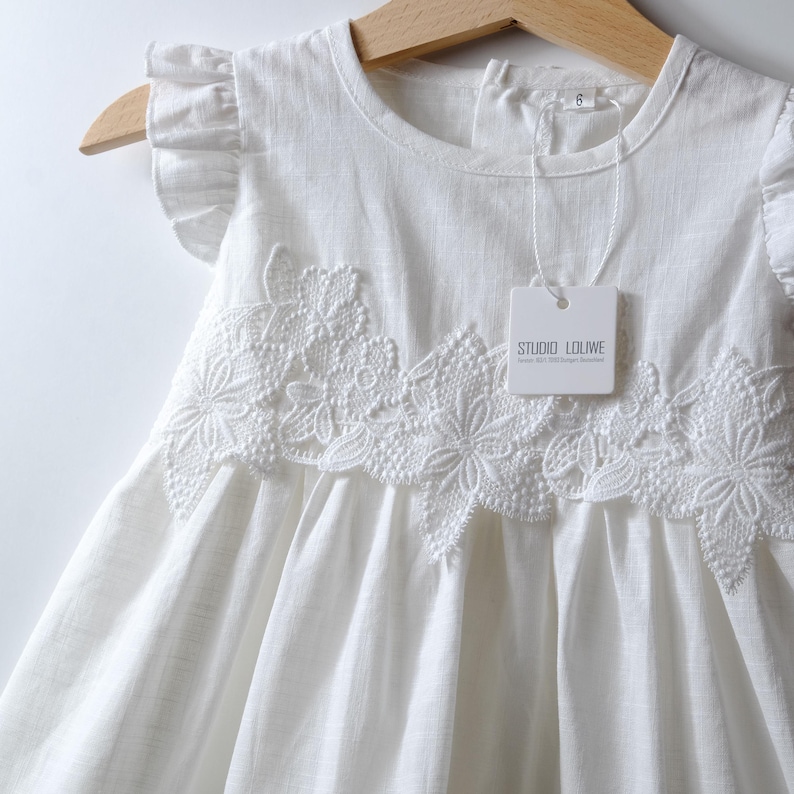 May include: A white sleeveless dress featuring ruffled cap sleeves, a round neckline, and a textured floral lace applique across the chest. A white tag attached to the garment displays the text STUDIO LOLIWE and the number 6 is visible on the collar.