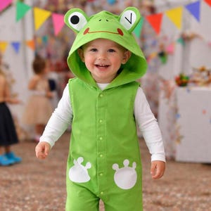 Puede incluir: Un niño con un disfraz de rana verde con capucha a juego que muestra ojos y una boca sonriente. El disfraz incluye una parte delantera abotonada y detalles de patas de rana blancas. El niño también lleva una camiseta blanca de manga larga.