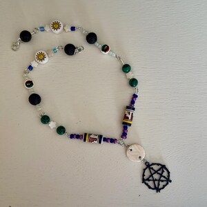 Puede incluir: Un collar hecho a mano con una variedad de cuentas en negro, verde, blanco y azul. El collar incluye cuentas en forma de sol, cuentas cilíndricas con un diseño y un colgante de pentagrama azul oscuro.