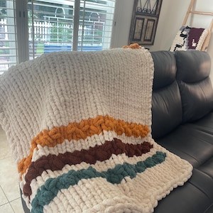 May include: A chunky knit throw blanket made of thick, cream-colored yarn. The blanket features three horizontal stripes in burnt orange, dark brown, and muted teal, creating a textured, soft appearance with large, visible loops throughout the design.