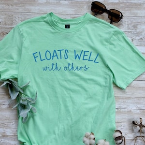 May include: A light green short-sleeved t-shirt featuring the blue text "FLOATS WELL with others" printed across the chest. The shirt has a crew neckline and is accompanied by a pair of dark-rimmed sunglasses positioned near the top right corner.