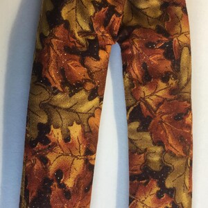 May include: A pair of doll pants featuring an elastic waistband and a fabric pattern of overlapping autumn leaves in shades of orange, brown, yellow, and dark green. The fabric has a subtle glittery finish throughout the leaf print design.