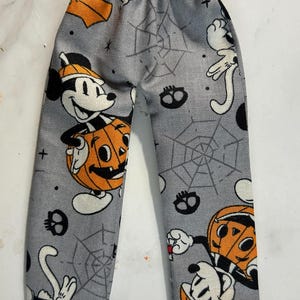 May include: Gray pants with an elastic waistband featuring a repeating Halloween print of Mickey Mouse characters wearing orange pumpkin costumes, black spiderwebs, and small black skulls scattered across the fabric in a festive pattern.
