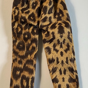 May include: A pair of miniature pants with a brown and tan leopard print pattern. The pants are made of a textured fabric and have an elastic waistband. The legs are straight and narrow.