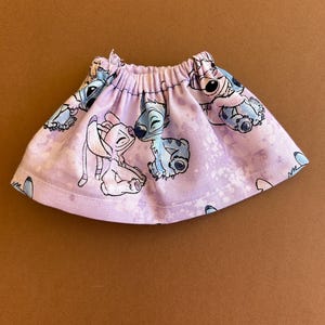 May include: A lavender skirt featuring a repeating pattern of cartoon characters in blue and white. The skirt has an elastic waistband and a flared shape. The fabric has a subtle, all-over white design.