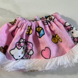 May include: A pink skirt with a white fluffy trim. The skirt is decorated with cartoon images of Hello Kitty, hearts, and other colorful shapes. The skirt is made of a soft, plush material.