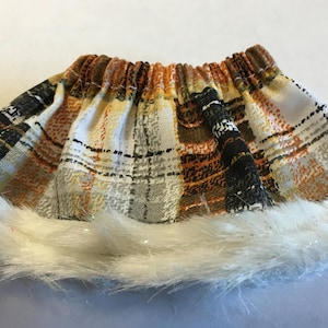 May include: A pleated skirt featuring a plaid pattern in shades of orange, black, white, and grey. The skirt has an elasticized waistband and is finished with a thick, white faux fur trim along the bottom hemline, creating a textured appearance.
