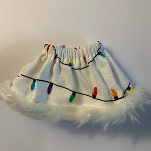 May include: A white skirt with a Christmas lights pattern. The skirt has a black outline of lights with colorful bulbs. The bottom edge is trimmed with white fluffy material.