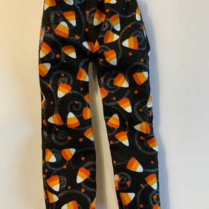 May include: A pair of doll pants featuring a black fabric base with a repeating pattern of candy corn in orange, yellow, and white, accented by small orange dots and swirling gray lines. The pants have an elasticized waistband at the top.