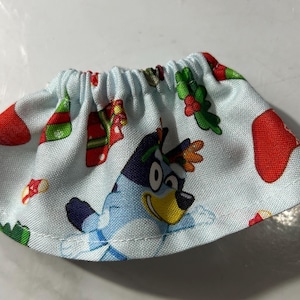 May include: A light blue skirt featuring a cartoon dog character and Christmas-themed patterns. The skirt has an elastic waistband and is decorated with red stockings, green holly, and other festive elements.