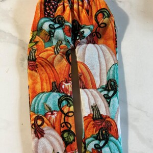 May include: A pair of doll pants featuring an all-over print of pumpkins and apples in shades of orange, teal, white, and green. The pants have an elasticized waistband and straight legs, showcasing a detailed harvest-themed fabric pattern.