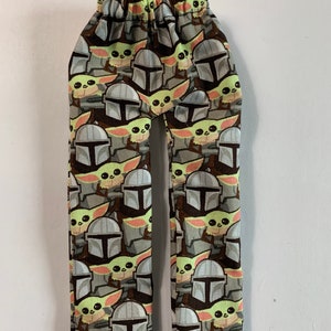 May include: A pair of patterned pants featuring a repeating design of a Mandalorian helmet and a green-skinned character. The pants have an elastic waistband and a brown, gray, and green color scheme.