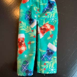 May include: Teal pajama pants with an elastic waistband. The pants feature a pattern of Christmas stockings with Mickey Mouse silhouettes, red berries, and snowflakes. The stockings have plaid patterns in red and blue. The pants are made of a soft fabric.