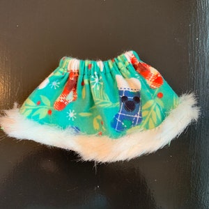 May include: A small, turquoise skirt with a white faux fur trim. The skirt features a Christmas-themed pattern with stockings, snowflakes, and Mickey Mouse silhouettes. The elastic waistband is gathered.