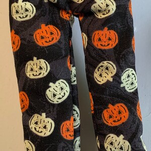 May include: Black pants with a repeating pattern of orange and white jack-o'-lanterns. The Halloween-themed pants are made of a dark fabric and feature a variety of smiling pumpkin faces, perfect for the spooky season.