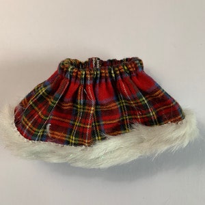May include: A miniature red plaid skirt with a white faux fur trim. The skirt features a classic tartan pattern in red, yellow, blue, and green. The elastic waistband is visible at the top.