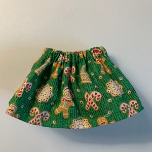 May include: A green skirt with a Christmas-themed print featuring gingerbread men, candy canes, and stars. The skirt has a gathered elastic waistband.