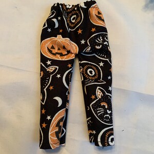 May include: Black fabric pants featuring a repeating Halloween pattern of orange jack-o'-lanterns, black cat faces, white crescent moons, and small stars. The pants have an elasticized waistband and a straight-leg cut with a simple hem.