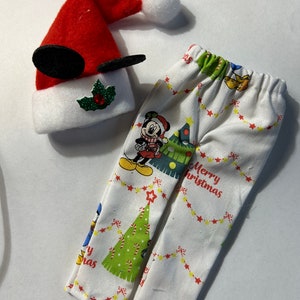 May include: A festive Christmas outfit for a doll, featuring a red Santa hat with Mickey Mouse ears and a white trim, and matching white pants printed with Mickey Mouse, Christmas trees, and the words "Merry Christmas".