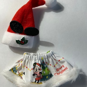 May include: A red Santa hat with black Mickey Mouse ears and a white pom-pom, next to a white skirt with a Mickey Mouse Christmas design and a white fur trim. The skirt has the words "Merry Christmas".