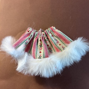 May include: A gathered skirt featuring a festive pattern of red and green stripes, small red hearts, and candy canes on a beige background. The hem of the skirt is trimmed with a thick, white faux fur border that extends around the entire bottom edge.