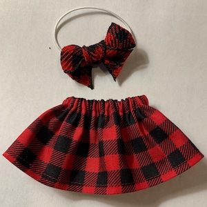 May include: A red and black plaid skirt and matching bow with a white elastic hair tie. The skirt is gathered at the waist. The bow is tied in a classic bow shape.