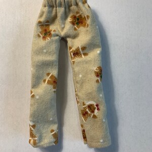May include: A pair of doll pants in a light beige fabric featuring a repeating pattern of brown gingerbread people with red and green accents and small white dots. The pants have a gathered elastic waistband and straight legs with hemmed cuffs.