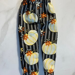 May include: A pair of doll pants featuring a vertical black and gray striped pattern. The fabric is decorated with a repeating print of light yellow pumpkins and small orange and white flowers. The pants have an elasticized waistband at the top.