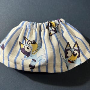 May include: A white and blue striped skirt with a blue dog character print. The dog is wearing a blue shirt and has a yellow face.