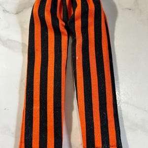 May include: A pair of pants featuring a vertical striped pattern in alternating black and bright orange colors. The garment has an elasticized waistband at the top and straight-cut legs that extend downward, constructed from a woven fabric material.