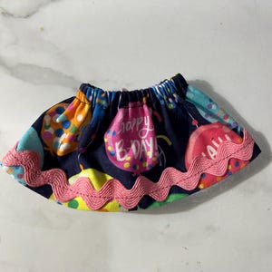 May include: A small, navy blue skirt with a colorful balloon print, including the words "Happy B-Day!" in pink. The skirt is trimmed with a pink, wavy ribbon. This festive skirt is perfect for a birthday celebration.