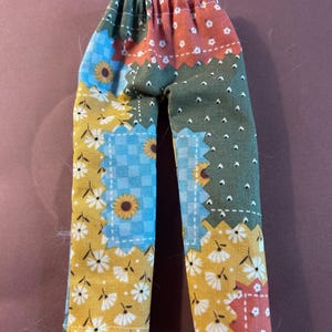 May include: Pants featuring a patchwork design with various fabric patterns including yellow sunflowers on a mustard background, blue and white checkered squares, and dark green fabric with small white dots, all finished with an elastic waistband.