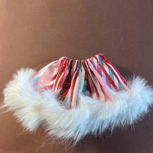 May include: A gathered fabric skirt featuring a festive pattern of red, white, and teal stripes with small snowflake accents. The hem of the skirt is trimmed with a thick, fluffy white faux fur border, creating a soft and textured appearance.