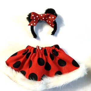 May include: A red skirt with large black polka dots and a white faux fur trim, paired with a black headband featuring two black fuzzy ears and a red bow with white polka dots. This set is designed for a child.