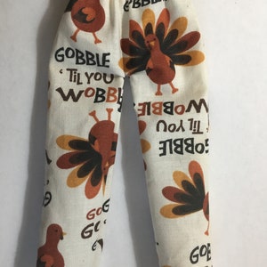 May include: Off-white pants featuring a repeating pattern of brown and orange turkeys and the text GOBBLE TIL YOU WOBBLE. The pants have an elasticized waistband and a straight-leg cut, showcasing a festive Thanksgiving-themed fabric design.
