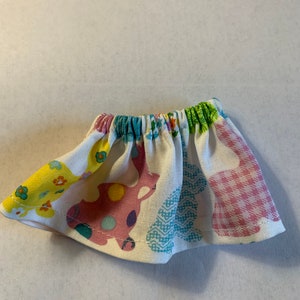 May include: A white fabric skirt with a colorful pattern of bunnies, flowers, and geometric shapes. The skirt has an elastic waistband.