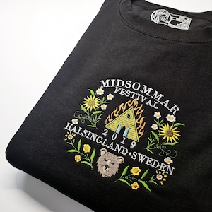 May include: Black sweatshirt with embroidered 'Midsommar Festival 2019 Helsingland Sweden' design. The design includes a burning house, flowers, and a bear. The sweatshirt is folded.