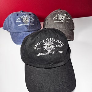 Puede incluir: Tres gorras de béisbol en azul, gris y negro. Cada gorra presenta el texto "SPOOKY ISLAND AMUSEMENT PARK" con un gráfico de calavera y el año "2002" bordado en blanco. Las gorras están hechas de tela suave.