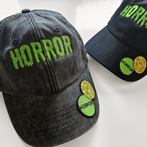 May include: Two black baseball caps with green embroidered text that says "HORROR". One cap has a yellow and green smiley face patch that says "BE KIND THESE ARE THE TIMES WE ARE IN". The other cap has a green patch that says "HORROR".