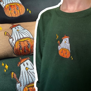 May include: A green sweatshirt with an embroidered design of a ghost holding a pumpkin. The ghost is wearing a hat and has a white sheet with a light blue trim. The pumpkin is orange with a green stem. There are small gold stars around the design.