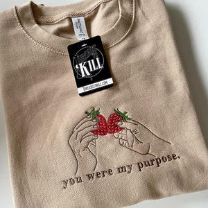 Peut inclure: T-shirt beige avec des fraises brodées et le texte "you were my purpose".