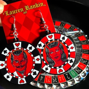 May include: A pair of red and black casino chip earrings featuring a devil and a devil woman design. The earrings are suspended from silver hooks.