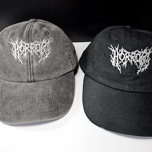 May include: Two baseball caps are shown, one in dark grey and the other in black. Both caps have the word "HORROR" embroidered in white, using a gothic font. The caps have a classic six-panel design with a curved peak.