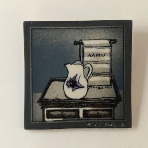 May include: Square ceramic tile with a painted still life scene. A white pitcher with a purple floral design sits on a small table. A towel with the word "AAMU" hangs above. The tile has a dark gray border.