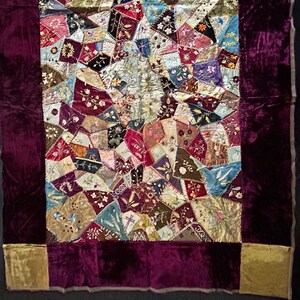 May include: A patchwork quilt with a burgundy velvet border and gold velvet corners. The quilt is made of many different fabrics and colours, including pink, blue, green, and brown. The quilt is decorated with embroidered flowers and other designs.