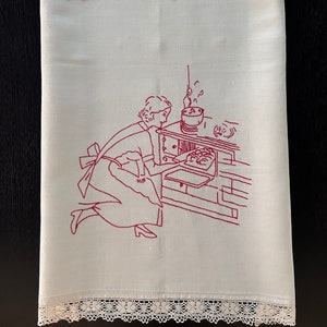 Op de afbeelding: Wit linnen keukendoek met een rood geborduurd ontwerp van een vrouw die bakt. De handdoek heeft een kantversiering en de initialen "J.K." zijn geborduurd in het midden bovenaan.