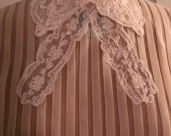 May include: A sheer, white blouse with vertical pleats and a delicate lace collar. The collar has a bow-like shape and features a floral pattern.