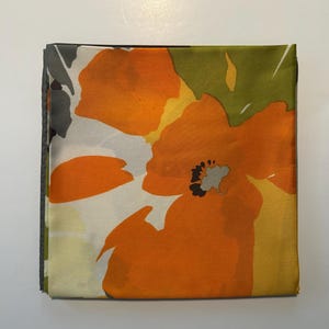 May include: Folded square fabric with a large orange floral pattern. The design features orange flowers with black centers, set against a background of white, yellow, and green. The edges are a dark gray.