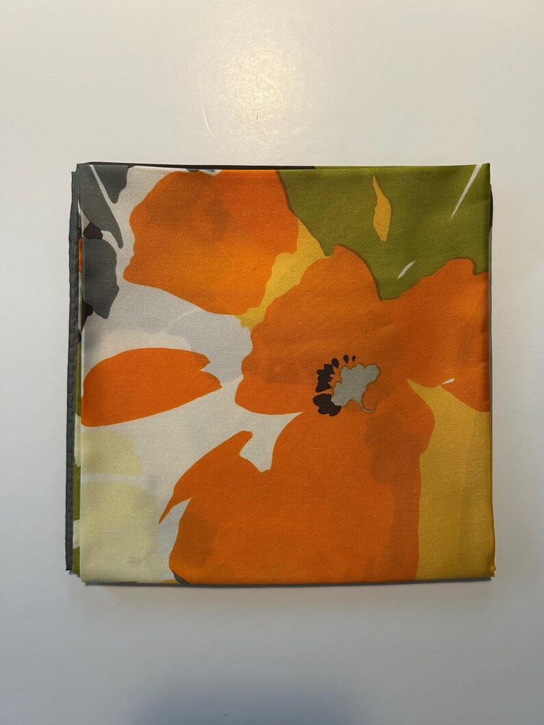 May include: Folded square fabric with a large orange floral pattern. The design features orange flowers with black centers, set against a background of white, yellow, and green. The edges are a dark gray.