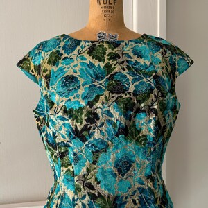 Puede incluir: Una blusa de color turquesa, verde y dorado con estampado floral y mangas cortas. La blusa es entallada y tiene un escote redondo.
